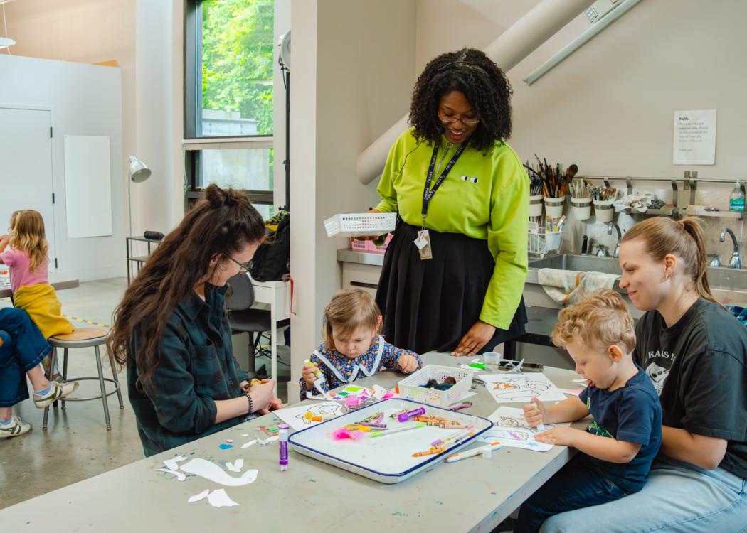 A pair of adults and children working on an art project while a teaching artist standing nearby smiles