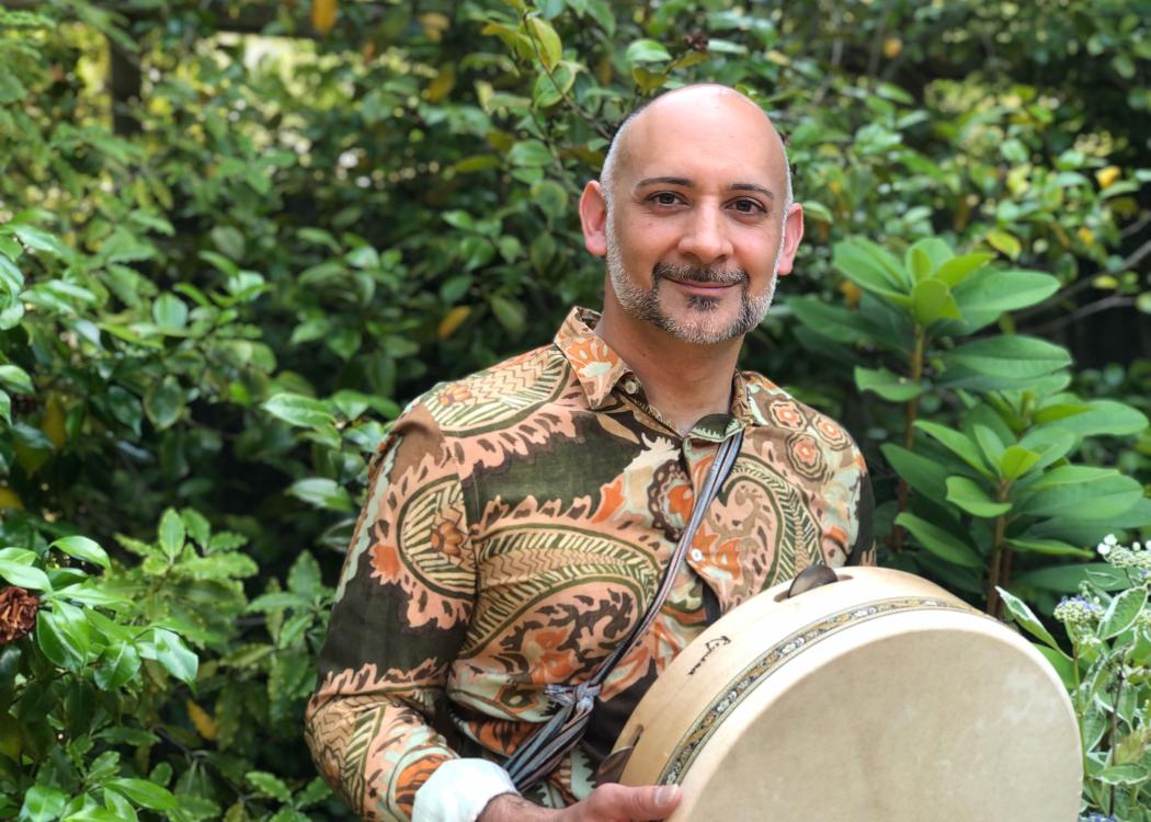 Musician with a long sleeve patterned shirt holding a drum and standing in front of a tree