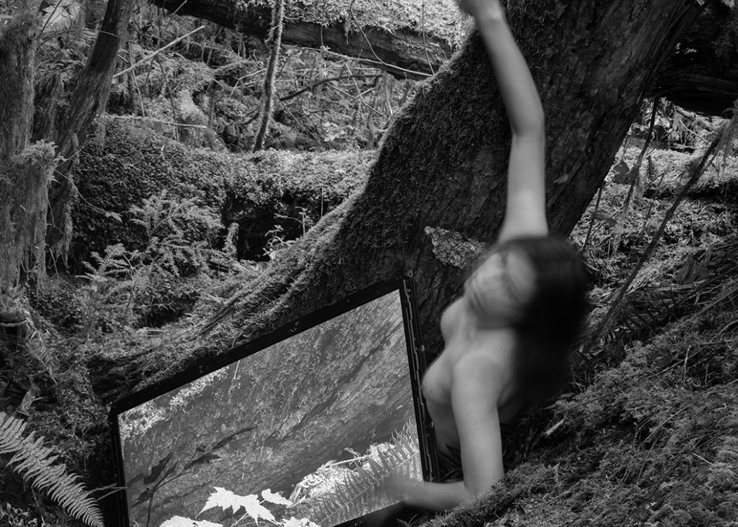 A black and white image of a nude human figure in a natural landscape with their body partially obscured by movement and a large LCD screen