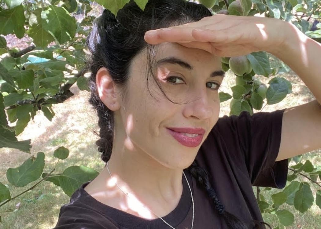 Photo of Ursula Brown, a woman with dark hair pulled back wearing a black t-shirt, surrounded by the leaves of a tree