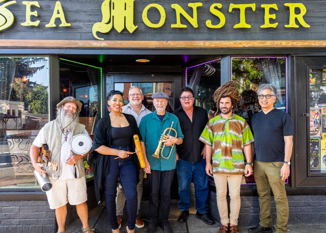 A diverse group of musicians posing outside a venue with a sign reading 'Sea Monster'