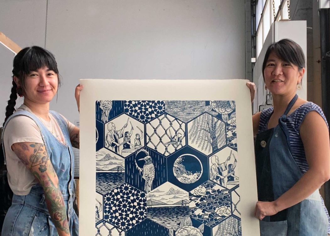 Artists Saiyare Refaei and Yoshi Nakagawa standing on either side holding their print "Sound", which is blue and white