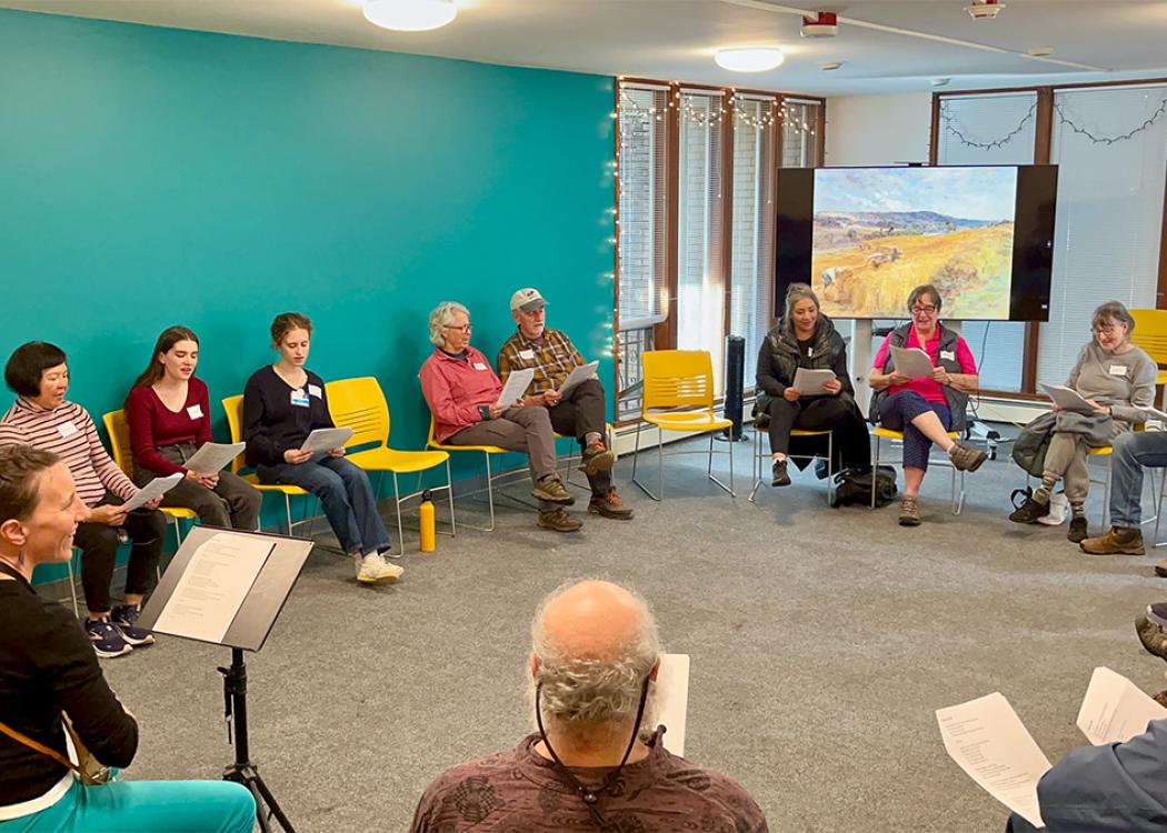 A group of about 15 people seated in a circle on chairs in a medium sized room. They are holding papers and following along as one person plays a guitar.