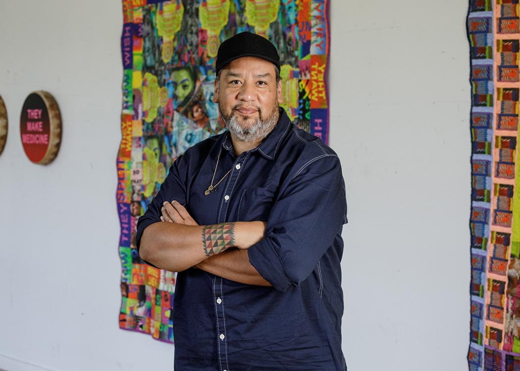 A person standing with their arms crossed in front of hanging textile artworks on a wall