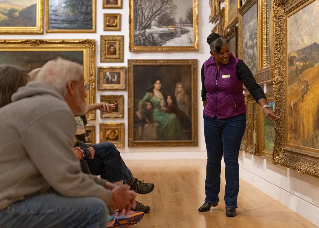 A person standing and pointing at an artwork in front of a small seated audience in a room filled with large artword in gold color frames