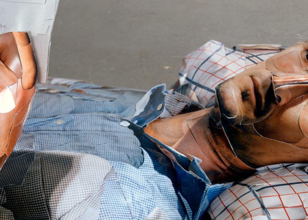 Detail of the face of a human figure made out of polystyrene and covered in prints to create the illusion of skin and clothing laying down with its knees up and reading from a piece of paper 