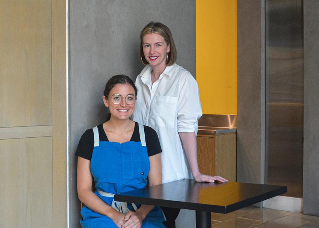 Two smiling women standing by a table, one sitting and one standing