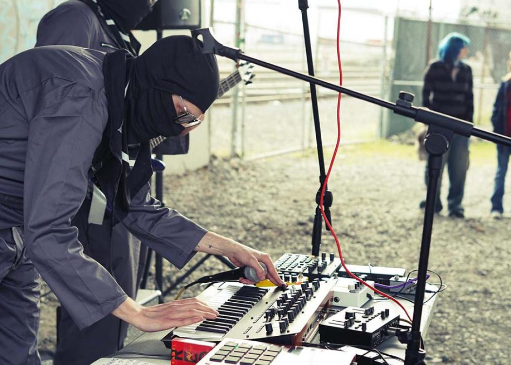 A person wearing a jumpsuit and a balaclava and holding a microphone adjusts electronic musical equipment on a table