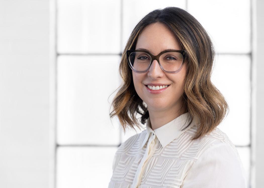 A professional headshot style photograph of a white woman with glasses wearing a white blouse