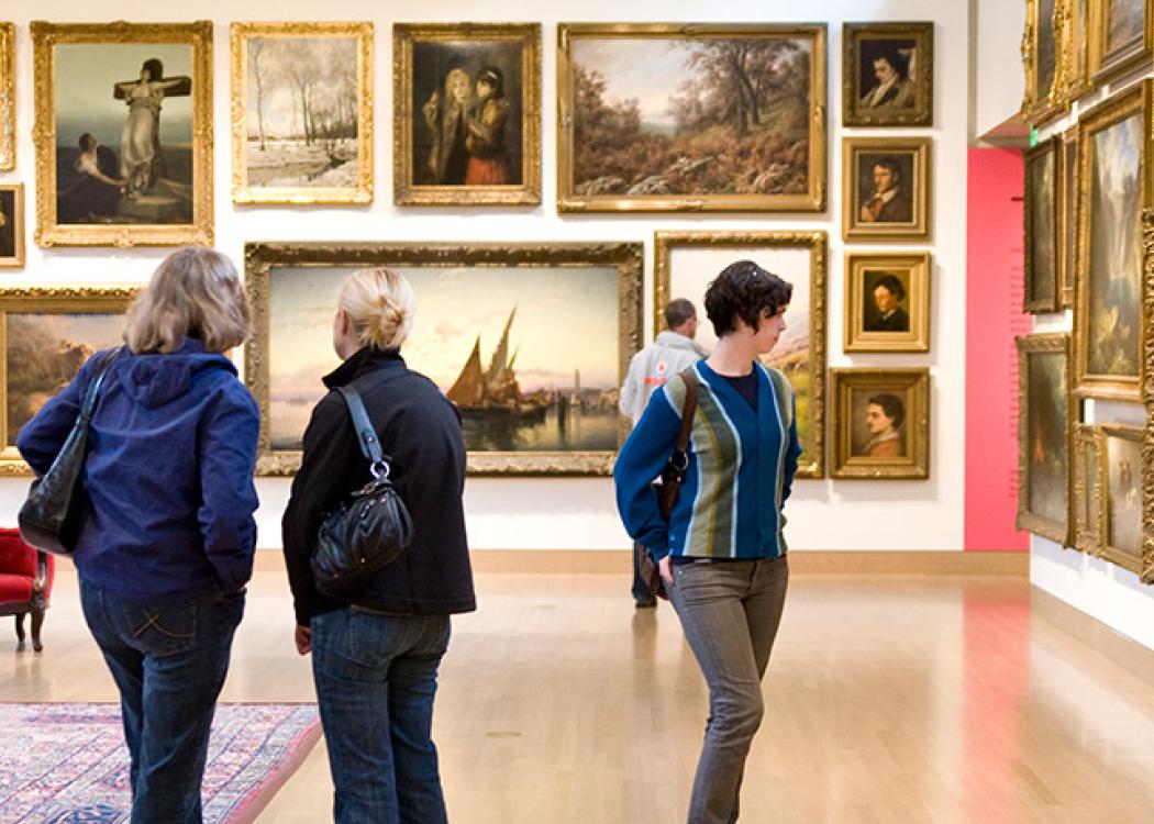 People in a large room hung with a carpet, chairs, and a potted plant with paintings hung floor to ceiling on the walls