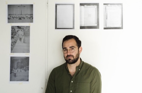 Man with dark hair and beard, dressed in a green button-down shirt, standing in his studio, surrounded by archival scans affixed to the wall