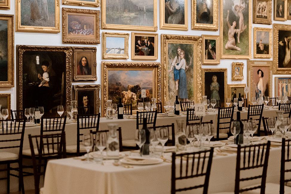 A beautifully set table is captured at the Frye Art Museum, featuring white tablecloths and glassware. The background showcases a gallery wall filled with framed artworks of various styles and subjects.