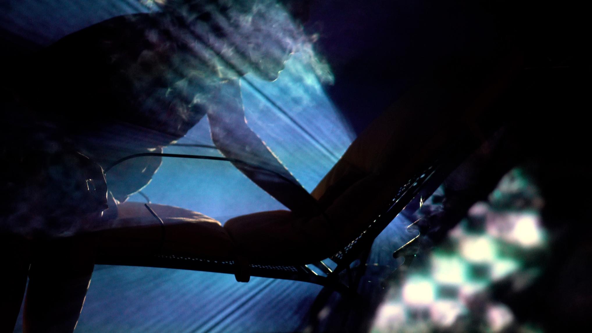 Video still of a dark figure in front of a blue wall with some light artifacts