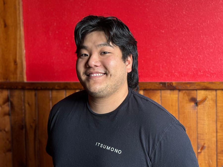 A smiling young man with dark hair is wearing a black shirt with the word "Itsumono" printed on it. He is standing in front of a wooden wall.