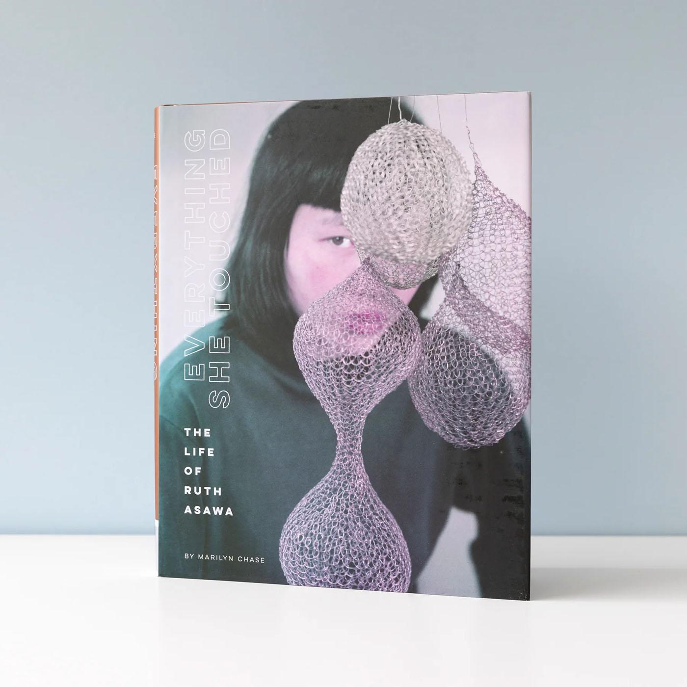 A book sitting on a white table. The cover has a woman's face slightly obscured by a hanging metal artwork.