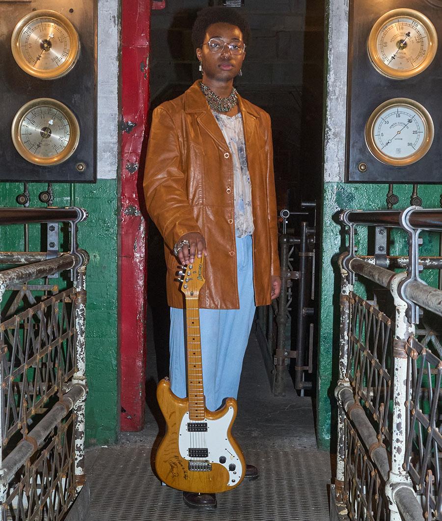 A younger black person in a brown leather jacket holding a guitar