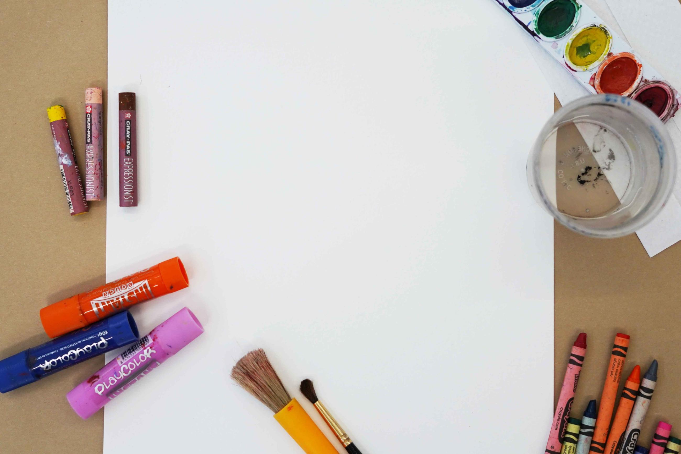 Photo of a blank sheet of paper with watercolors, pastels, crayons, and other art supplies around it