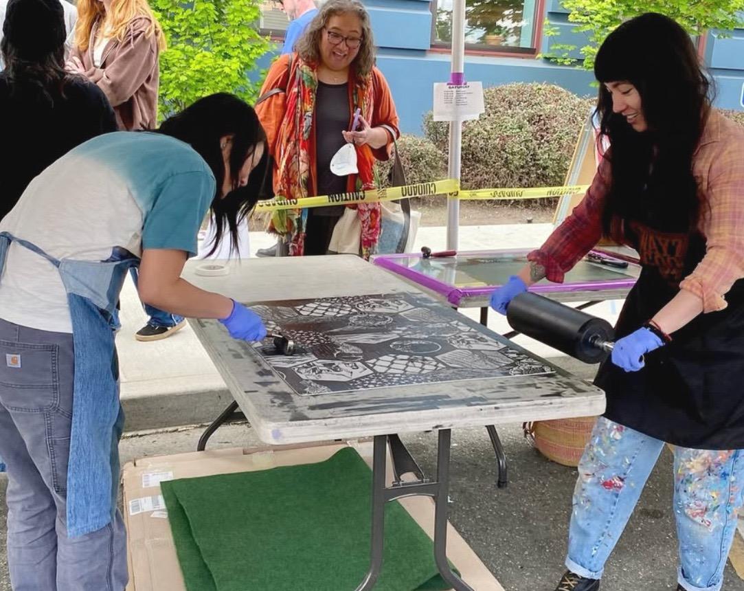 Artists Saiyare Refaei and Yoshi Nakagawa standing on either side of a table inking the linocut for "Sound"