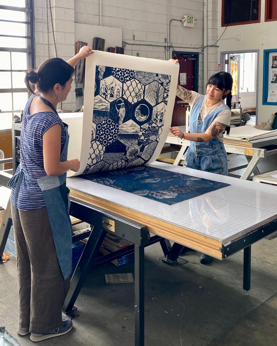 Artists Saiyare Refaei and Yoshi Nakagawa standing on either side of a table lifting their print "Sound", which is blue and white, in an art studio
