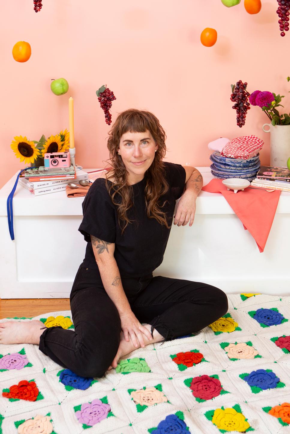 Cece Edson wearing all black while sitting on a colorful quilt with flowers, fruit, and tableware on a low shelf behind her