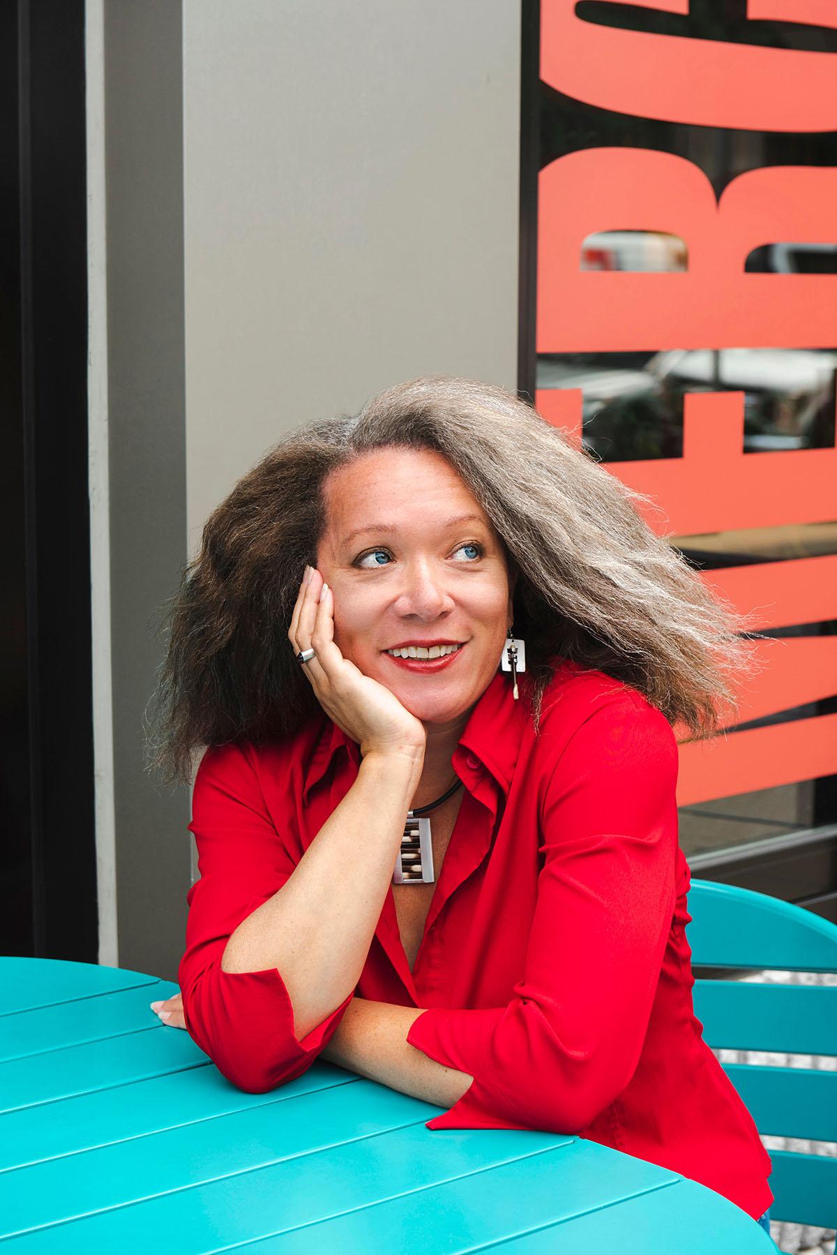 A woman in a bright red shirt sitting at a blue table, leaning her face on her hand and smiling
