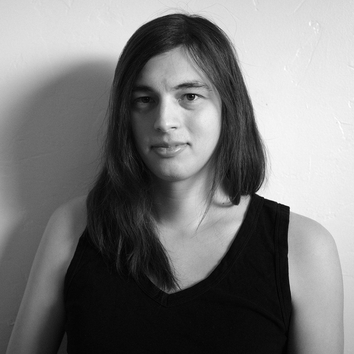 A tightly cropped black and white portrait photo of a woman with long dark hair against a white wall