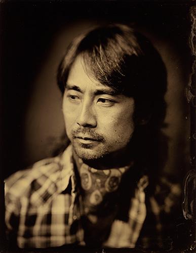 A black and white photograph of a man with shoulder-length dark hair wearing a flannel shirt and a bandana around his neck