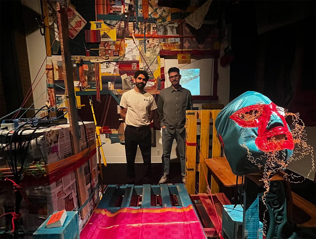 Two people standing in a room in front of lighting equipment and posters. There is a nacho libre Mexican wrestling mask handing on a post in the foreground