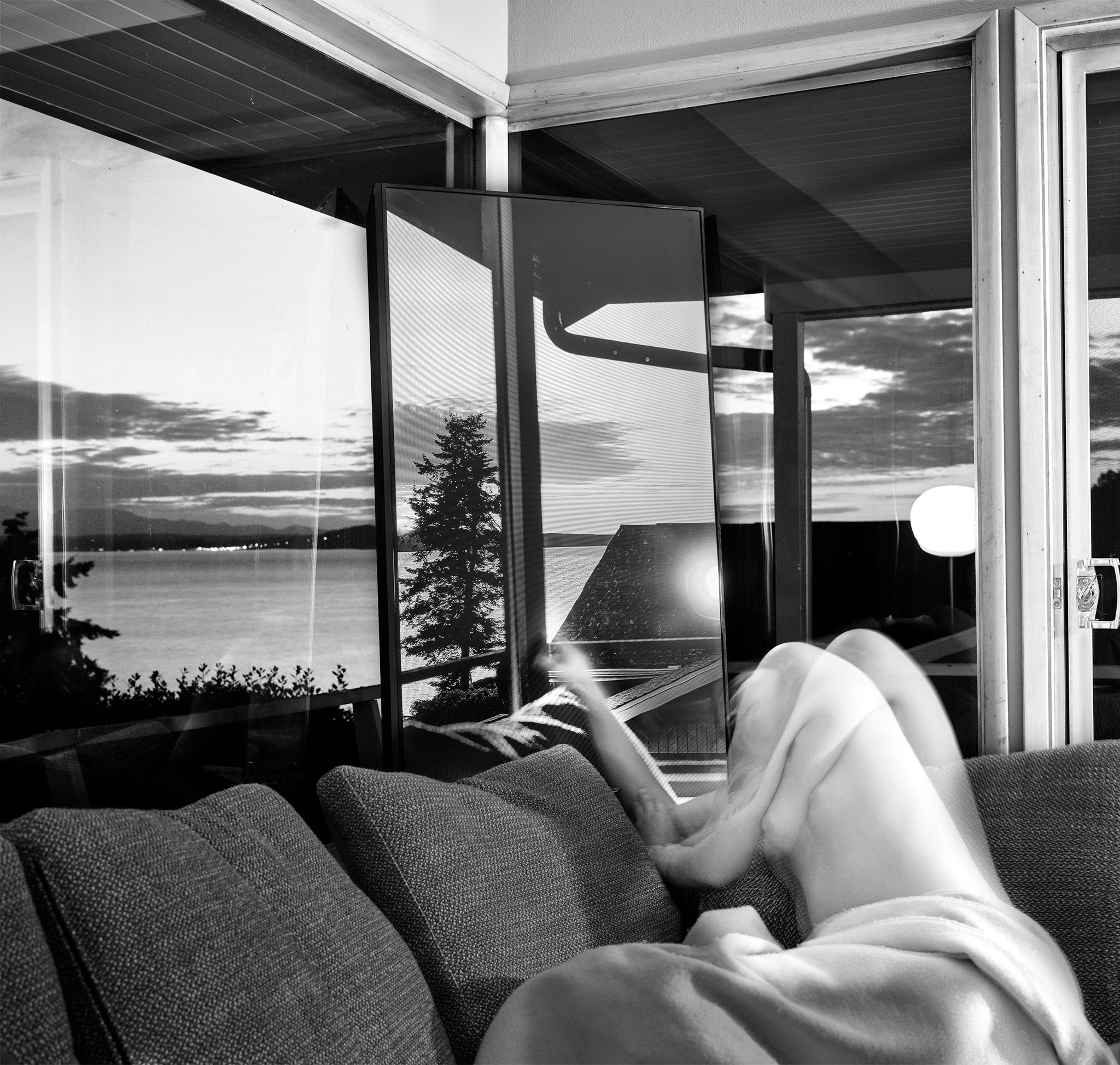 Photograph of a ghostly figure on a couch near a set of windows and a screen looking out at a body of water